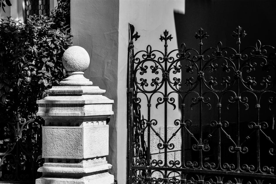 Black and white image of a decorative iron gate and stone post under bright sunlight, showcasing intricate details.